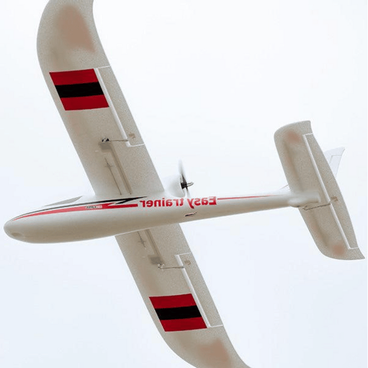 White remote control model airplane with red and black stripes on the wings and "Easy Trainer" written on the side, flying with a spinning propeller, viewed from below.