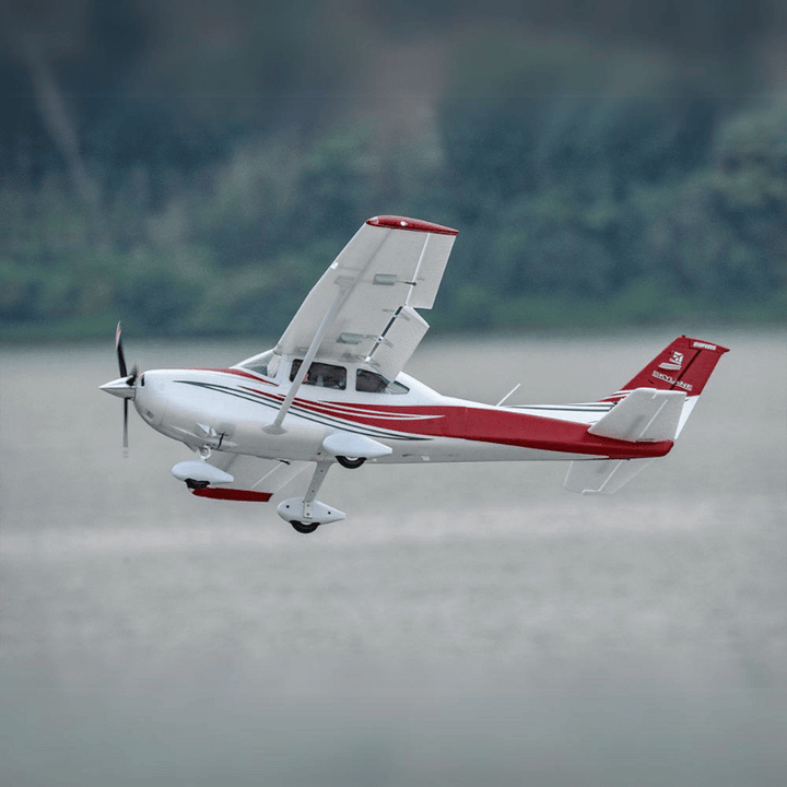 White and red single-engine small airplane flying over a body of water with blurred green trees in the background.