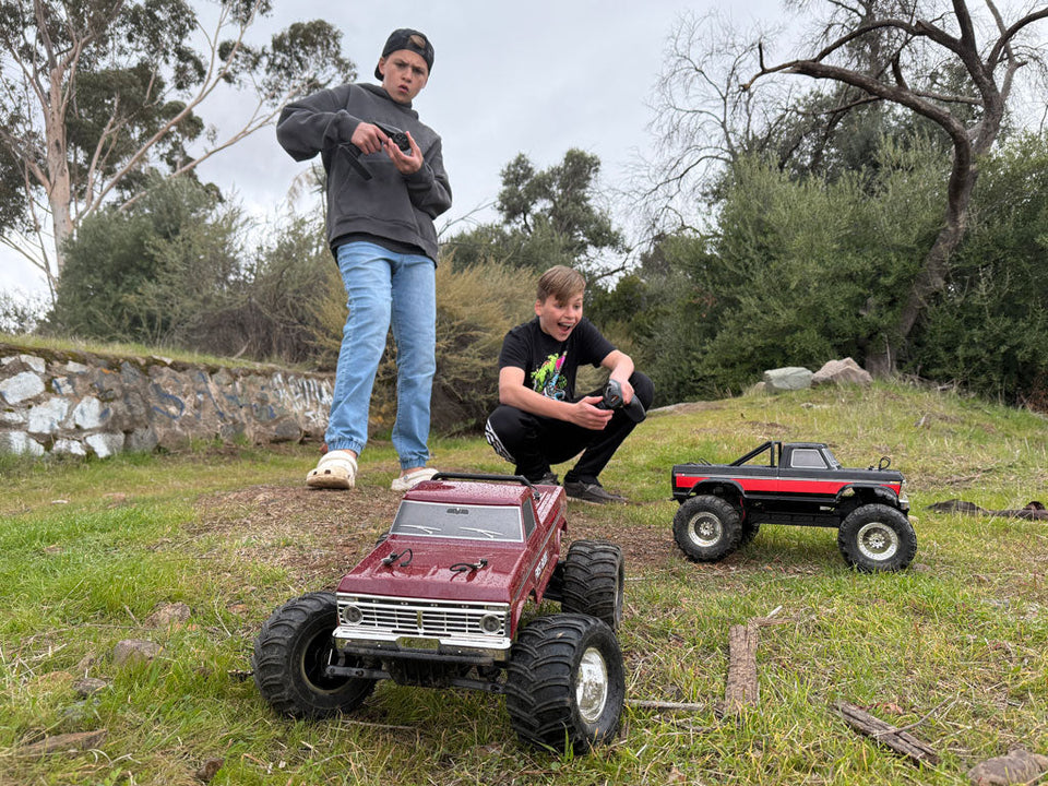 Two boys hold controllers while driving two Fmshobby RC trucks on a grassy hill
