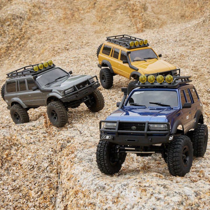 Three detailed Fmshobby RC Trucks navigate a rugged rocky terrain, highlighting their realistic off-road design