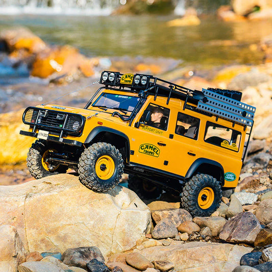 Yellow RC Land Rover Defender climbing rocks near a stream, featuring Camel Trophy decals and rugged off-road details.