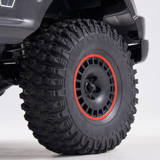 A close-up view of a rugged off-road tire with deep tread patterns, mounted on a black wheel rim featuring a red accent ring. The background is neutral and minimalistic.