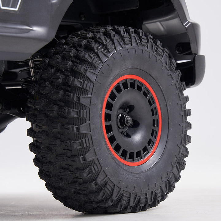 A close-up view of a rugged off-road tire with deep tread patterns, mounted on a black wheel rim featuring a red accent ring. The background is neutral and minimalistic