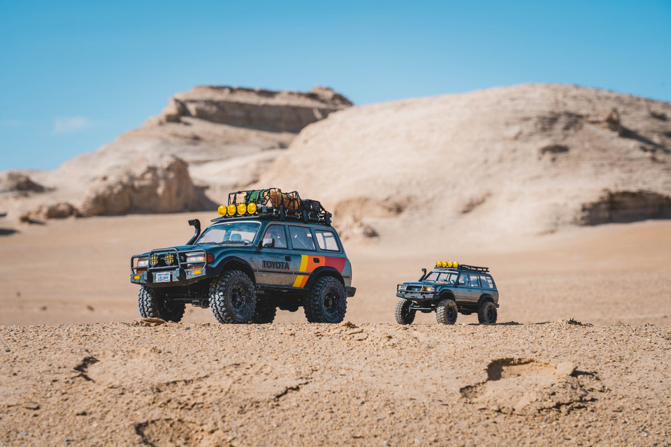Two Fmshobby Toyota RC crawlers sit on a sandy desert ridge with hills in the background