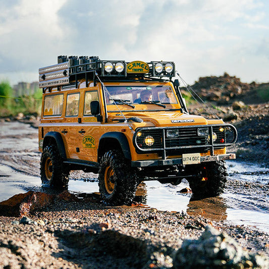 A yellow Land Rover Defender off-road vehicle with Camel Trophy branding, equipped with roof lights, a roof rack, and off-road tires, driving on a muddy trail with a backdrop of wasteland and blue sky.