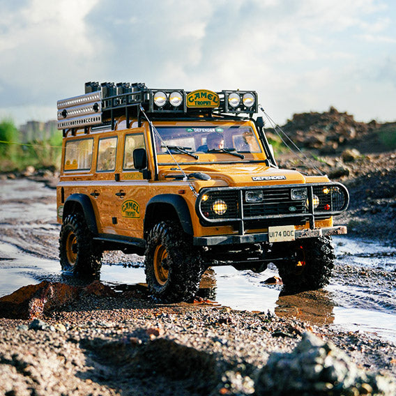 A yellow Land Rover Defender off-road vehicle with Camel Trophy branding, equipped with roof lights, a roof rack, and off-road tires, driving on a muddy trail with a backdrop of wasteland and blue sky.