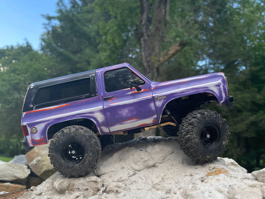 This image depicts a custom-painted, off-road RC truck with a distinctive purple and orange color scheme, positioned on a rocky outcrop, showcasing its rugged, adventurous capabilities.