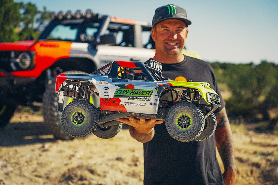 Man holding a remote-controlled truck in an outdoor setting with another truck in the background.