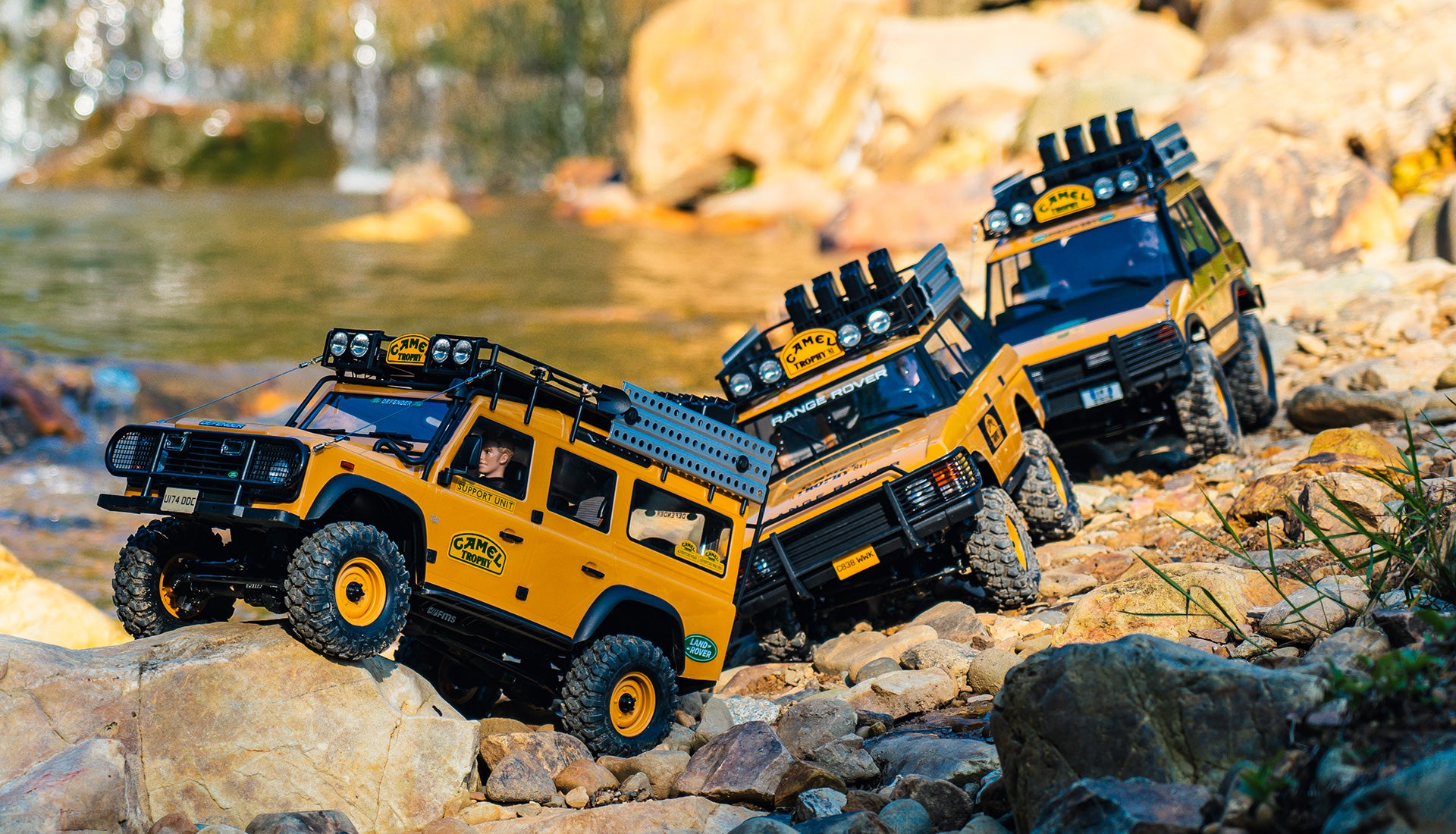 Three modified off-road Camel Trophy Land Rovers navigating a rocky, forested environment.