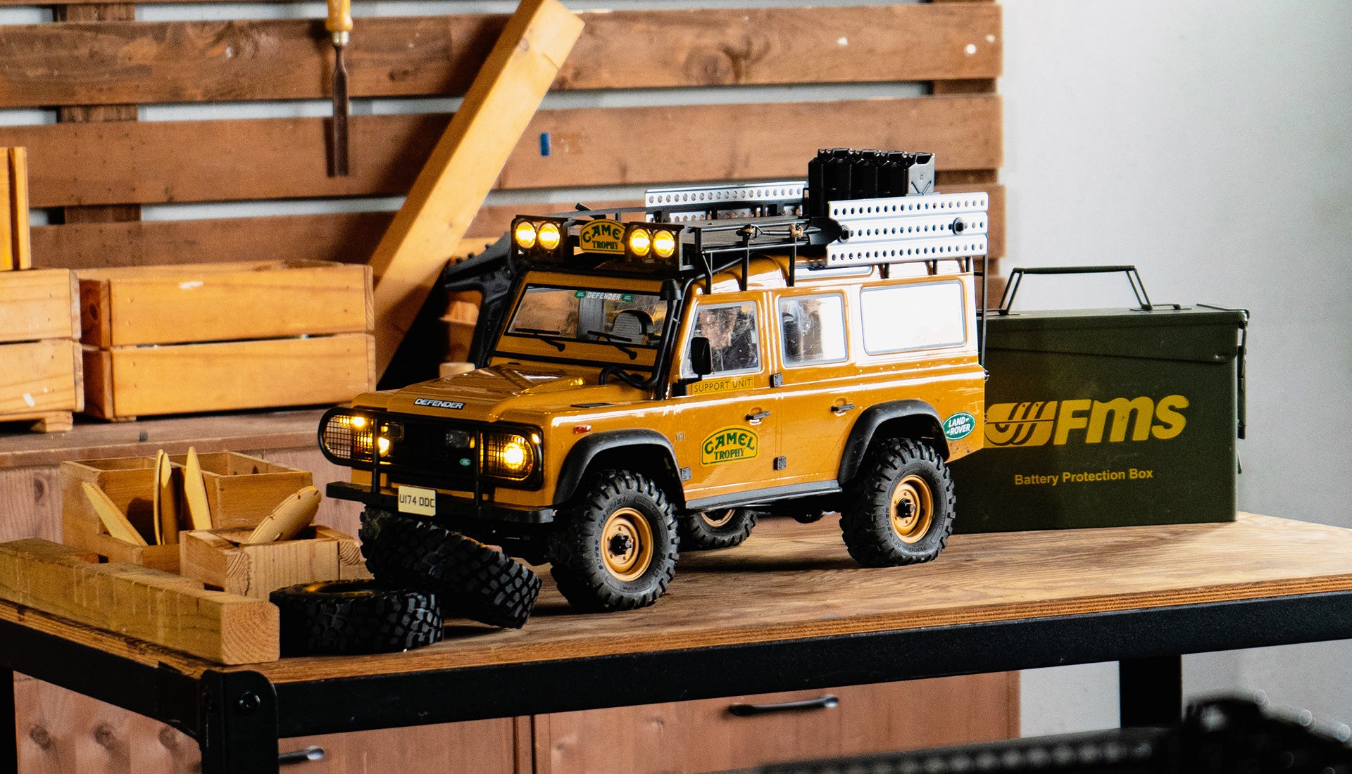 The image shows a highly modified off-road Camel Trophy edition Land Rover vehicle on a wooden shelf, with a battery protection box next to it. The vehicle is equipped with additional lighting and accessories to support its extreme adventure capabilities.
