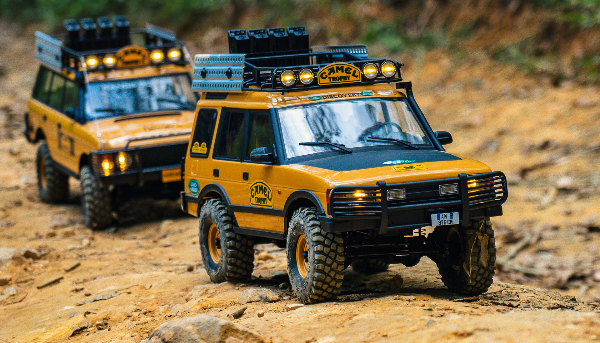 Two modified Camel Trophy Land Rovers traversing a rugged, wooded terrain.