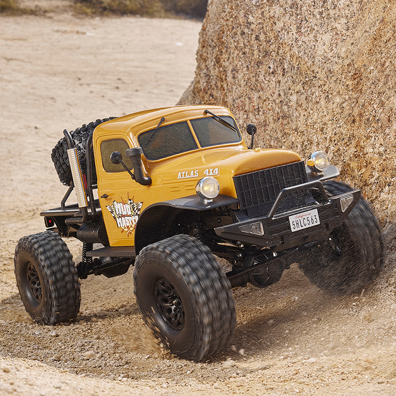 Yellow ROCHOBBY 1:10 Atlas 4x4 Off-Road Truck RS climbing rock on sandy terrain with large tires and 'MUD MASTER' decal.